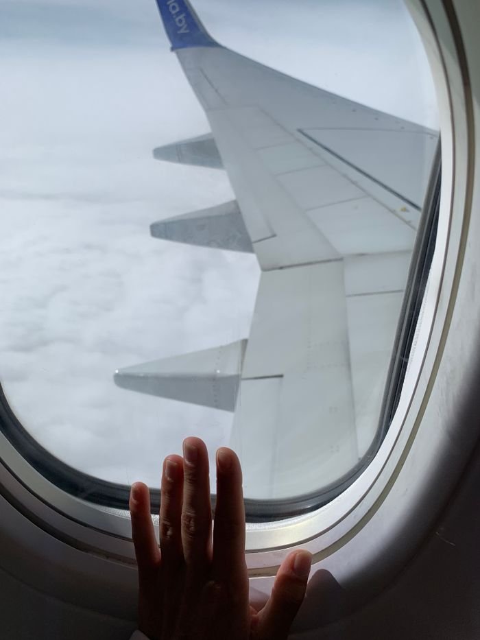 View from an airplane window showing a hand and the aircraft wing above the clouds.